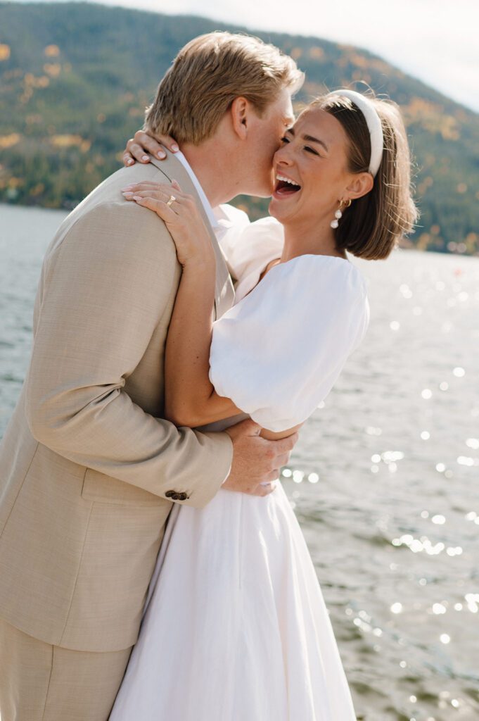 A bride and groom laughing together by a lake, demonstrating why you should select a wedding photographer whose documentary-style captures your true personality.