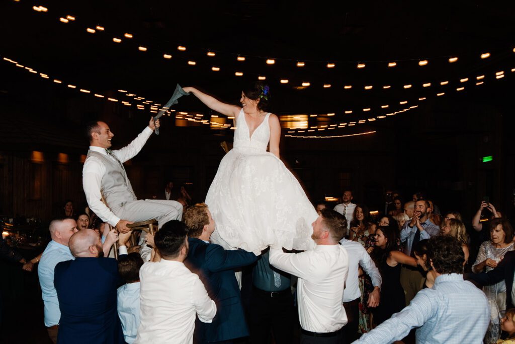 A high-energy candid photo of a bride and groom being lifted on chairs during a traditional wedding reception, showing how to select a wedding photographer who captures movement and joy.