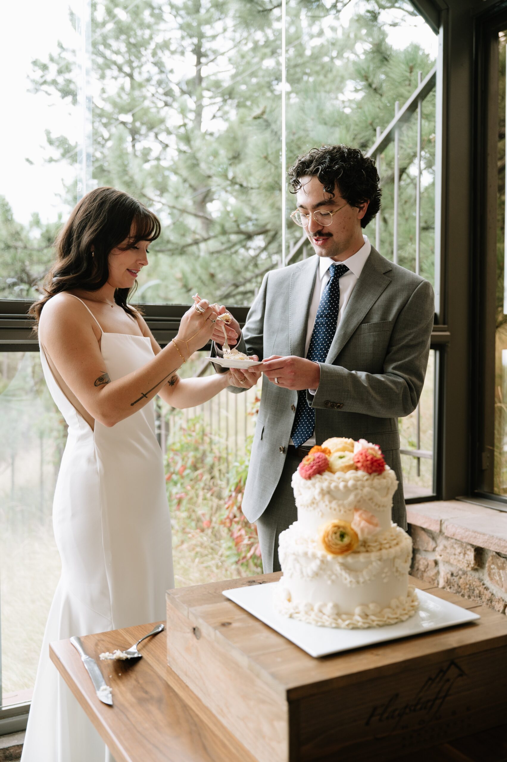 Bride feeding groom a bite of cake by a window overlooking trees, capturing a relaxed reception during the best month to get married in Colorado