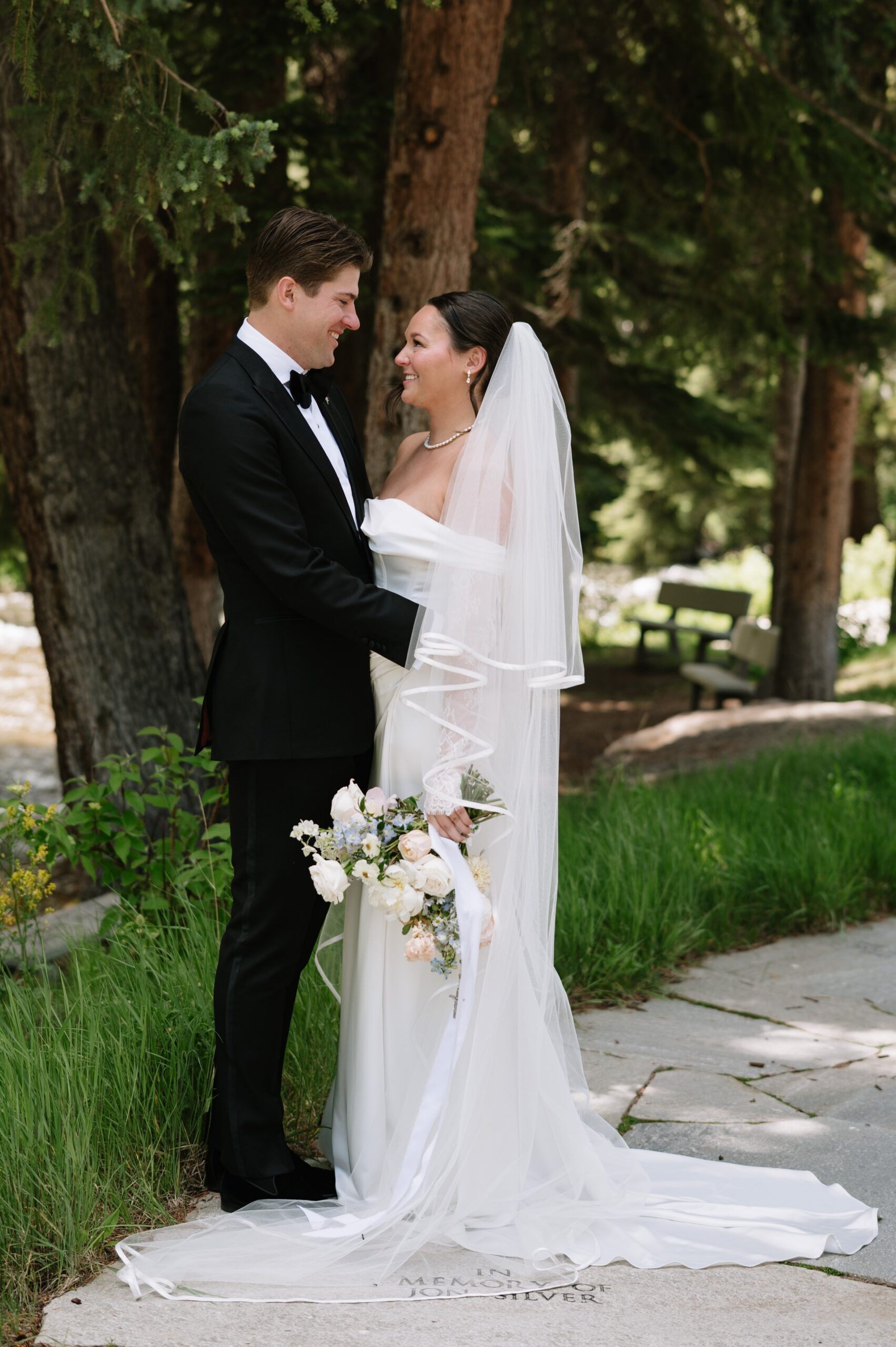Elegant bride and groom embracing beneath tall pine trees, reflecting how summer is often the best time of year to get married in Colorado for shaded outdoor portraits.