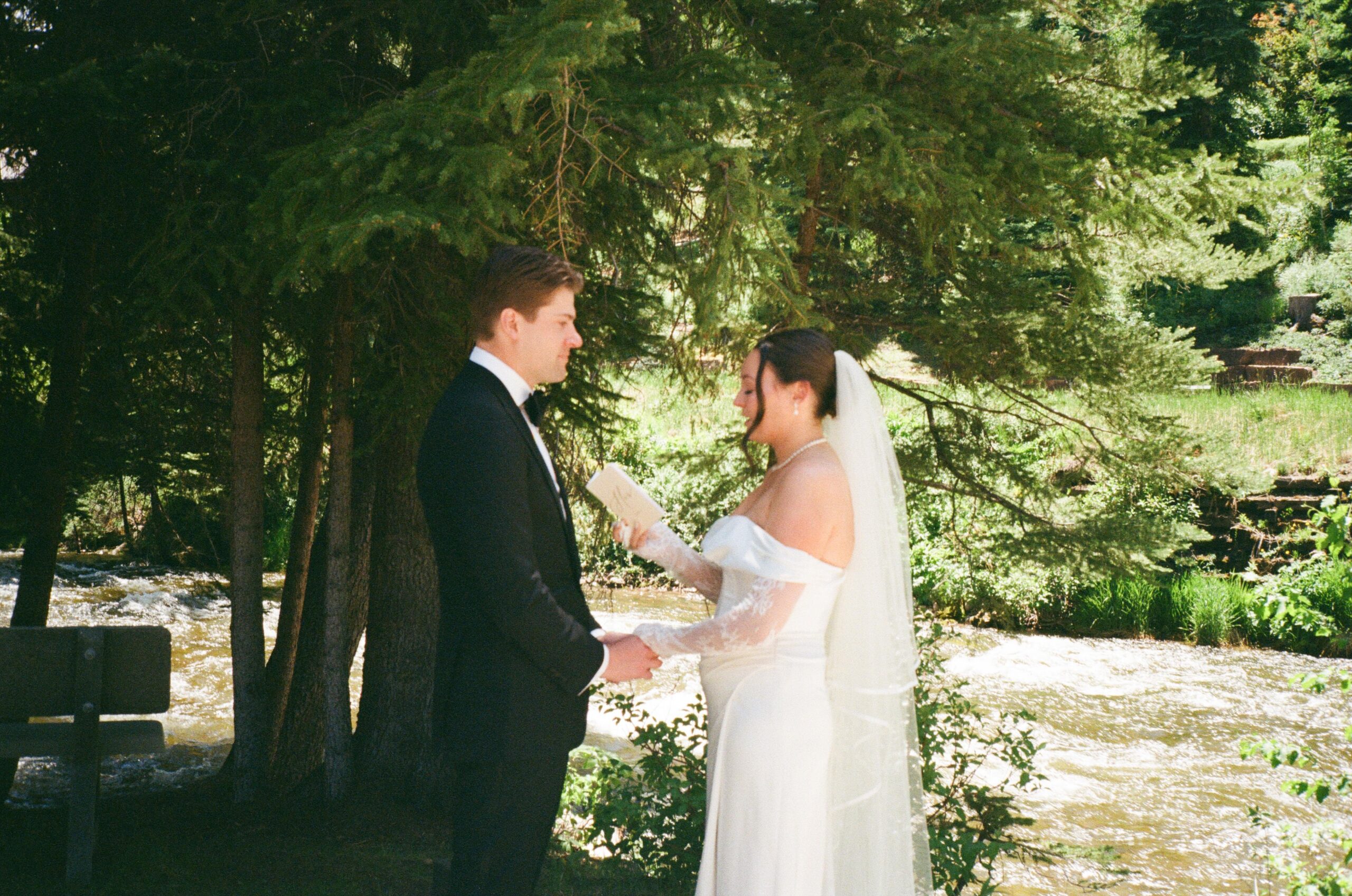 Bride reading vows to groom beside a flowing river under tall trees, a peaceful setting that shows why summer is often the best time to get married in Colorado.