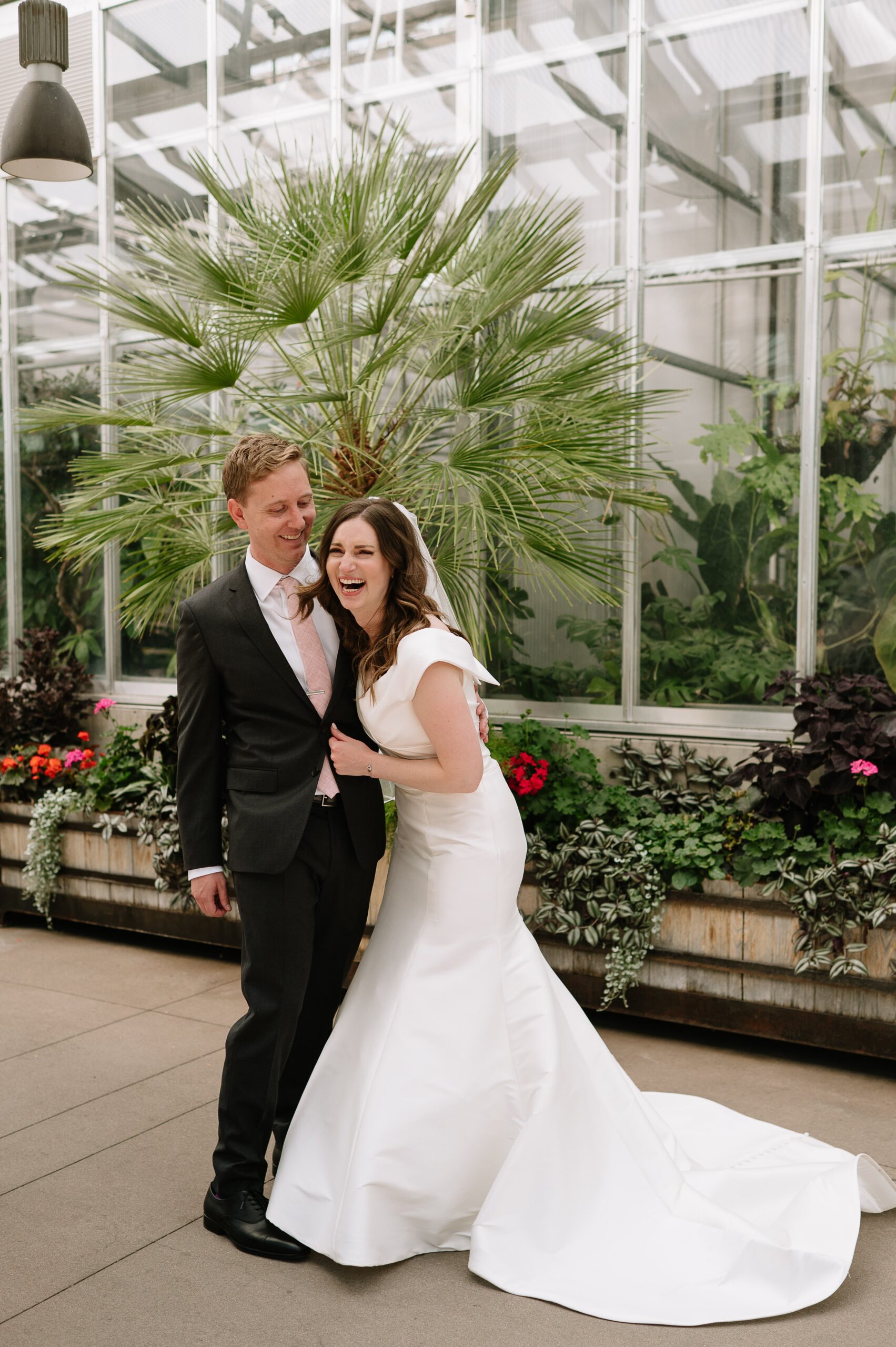 Joyful couple sharing a candid moment during their botanic gardens Denver wedding, standing in front of lush greenery and a glass greenhouse.