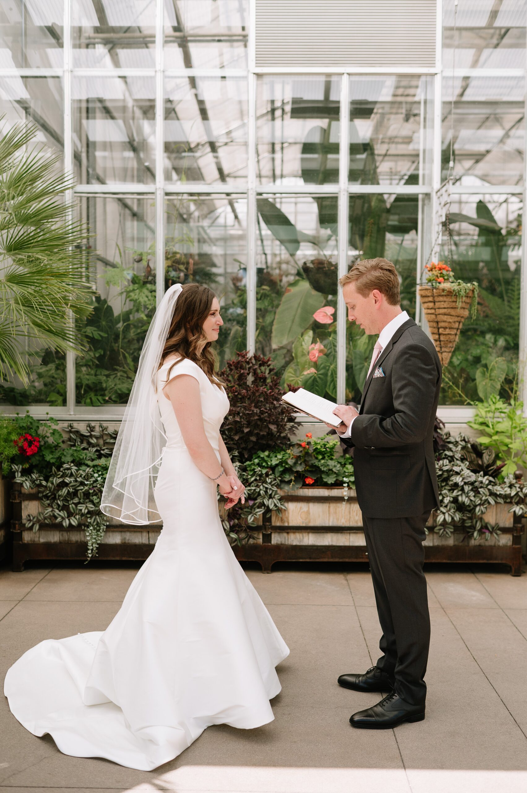 Bride and groom exchanging vows during a wedding at Denver Botanic Gardens, surrounded by vibrant plants and a bright conservatory backdrop.