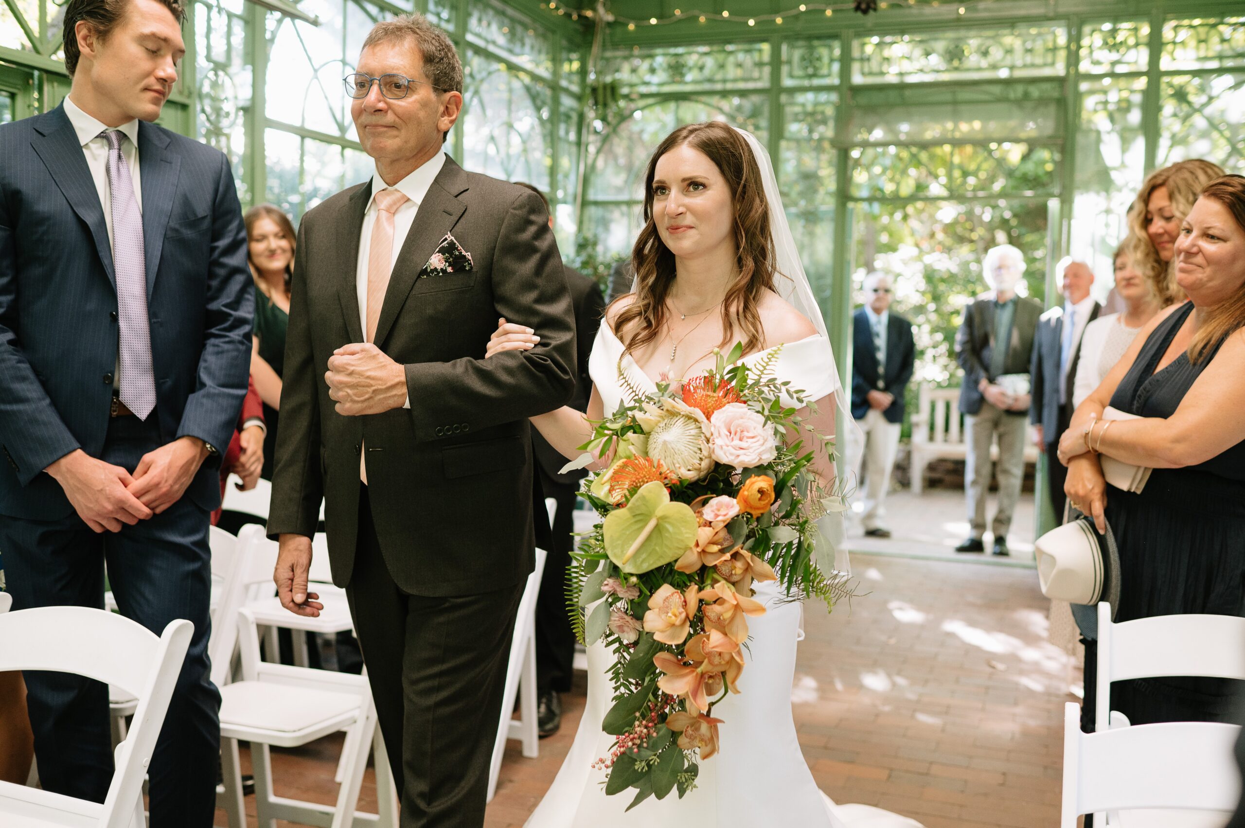 Bride walking down the aisle with her father during a Denver Botanic Gardens wedding, holding a colorful bouquet as guests look on inside the garden conservatory.