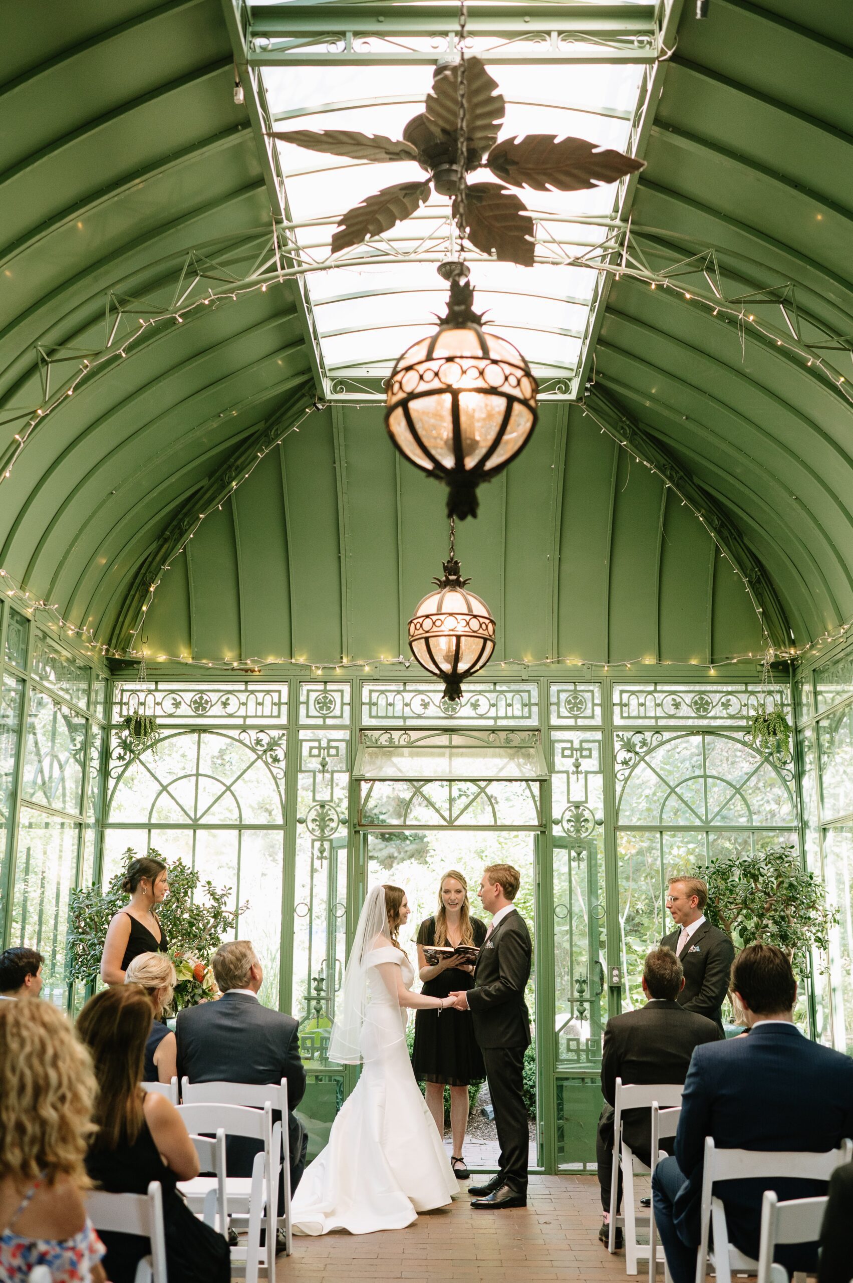 Wide view of a botanic gardens denver wedding ceremony inside a glass conservatory, with the couple exchanging vows beneath elegant green architecture and soft lighting.