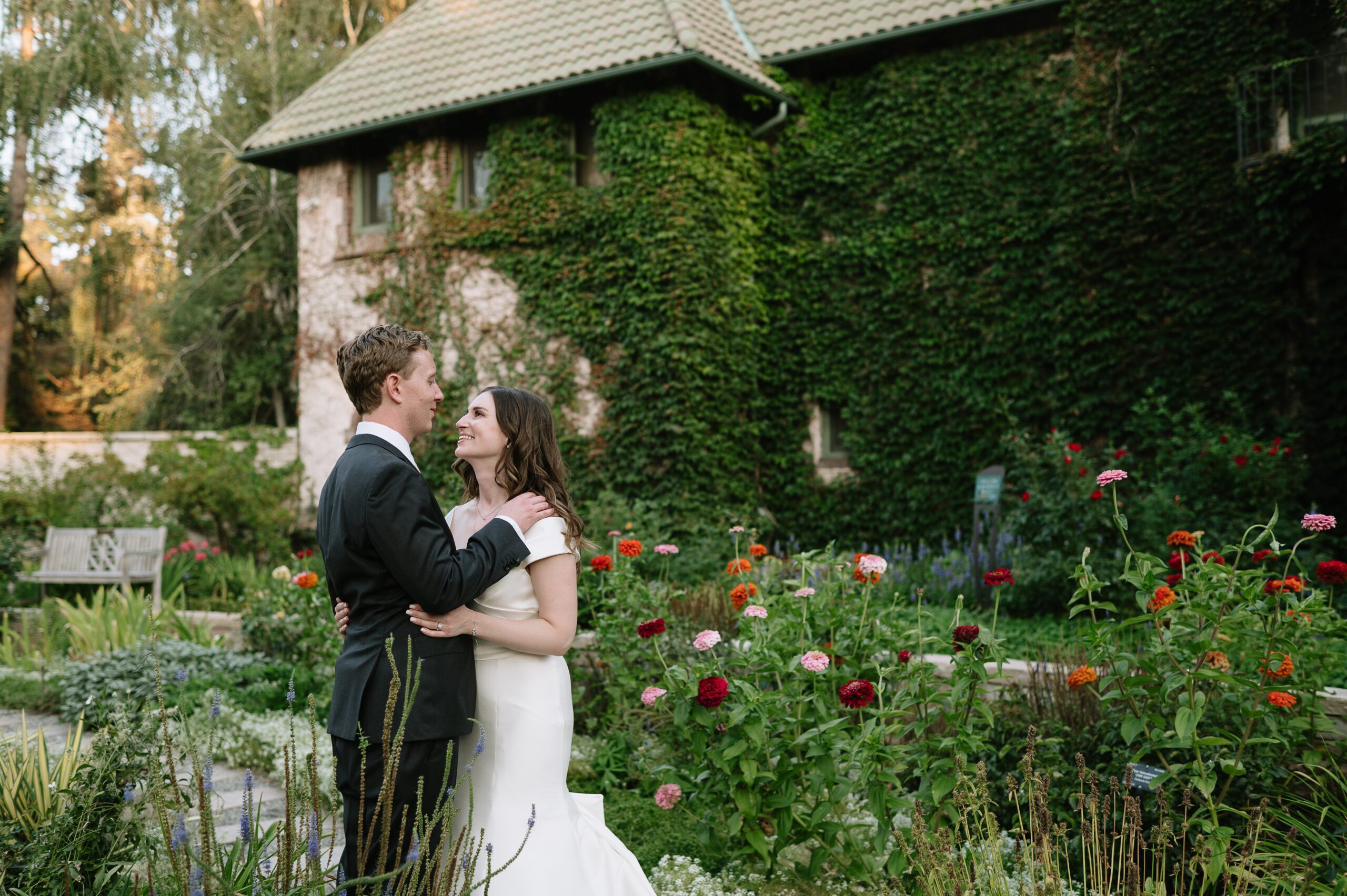 Bride and groom embracing in a lush flower garden during a denver botanic gardens wedding, with colorful blooms and an ivy-covered building in the background.