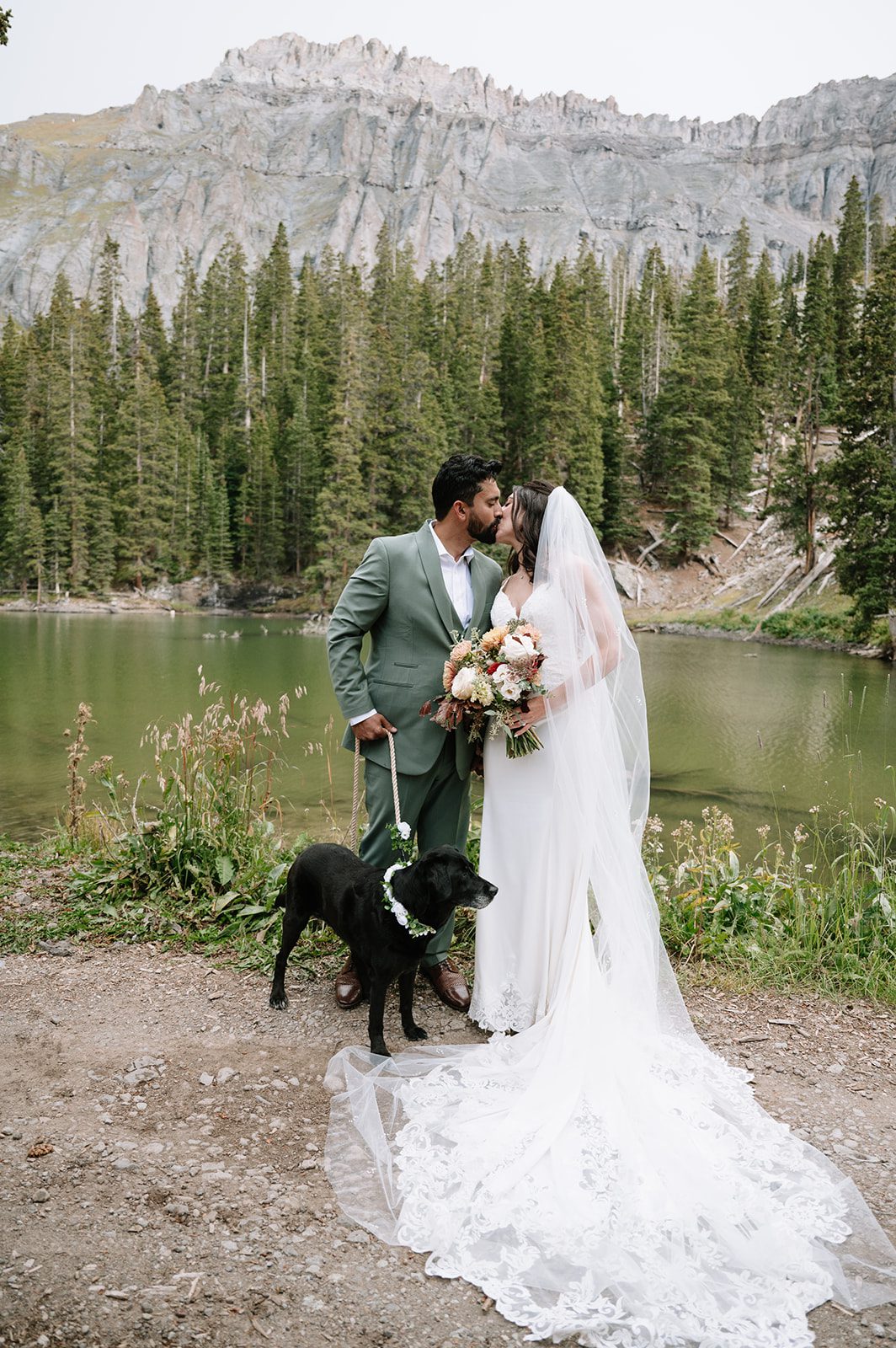 Newlyweds sharing a kiss by a mountain lake with their dog at a scenic small wedding venue in Colorado. 