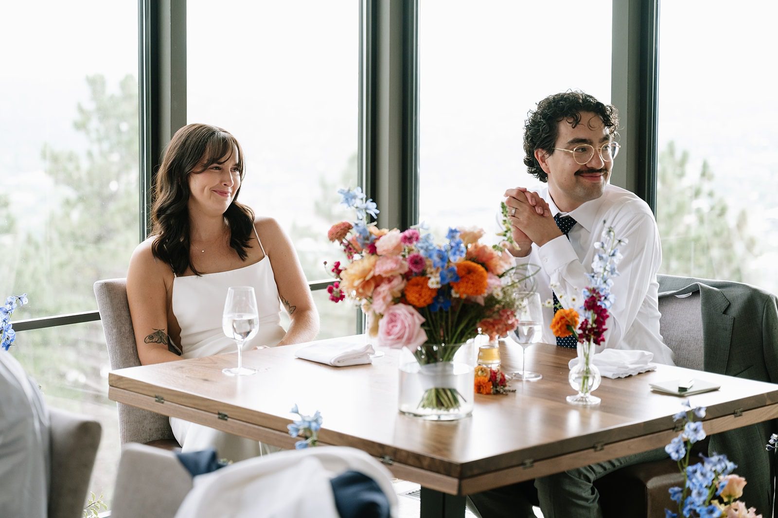 Bride and groom sit at a wooden table beside a colorful floral centerpiece during their brunch wedding reception.