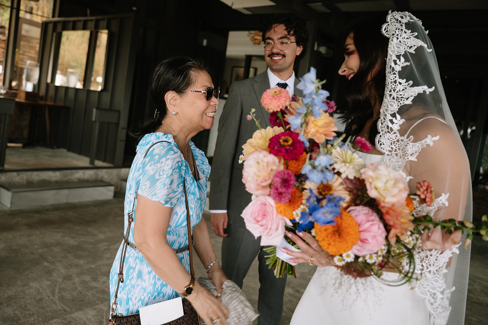 Bride holding a colorful bouquet smiles while greeting a wedding guest as the groom stands behind them.