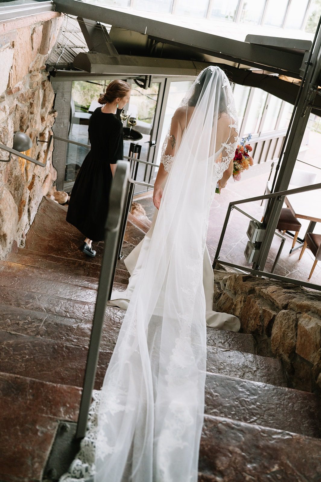 Bride descends a stone staircase inside Flagstaff House with her lace-edged veil trailing behind her as a woman in black walks ahead.