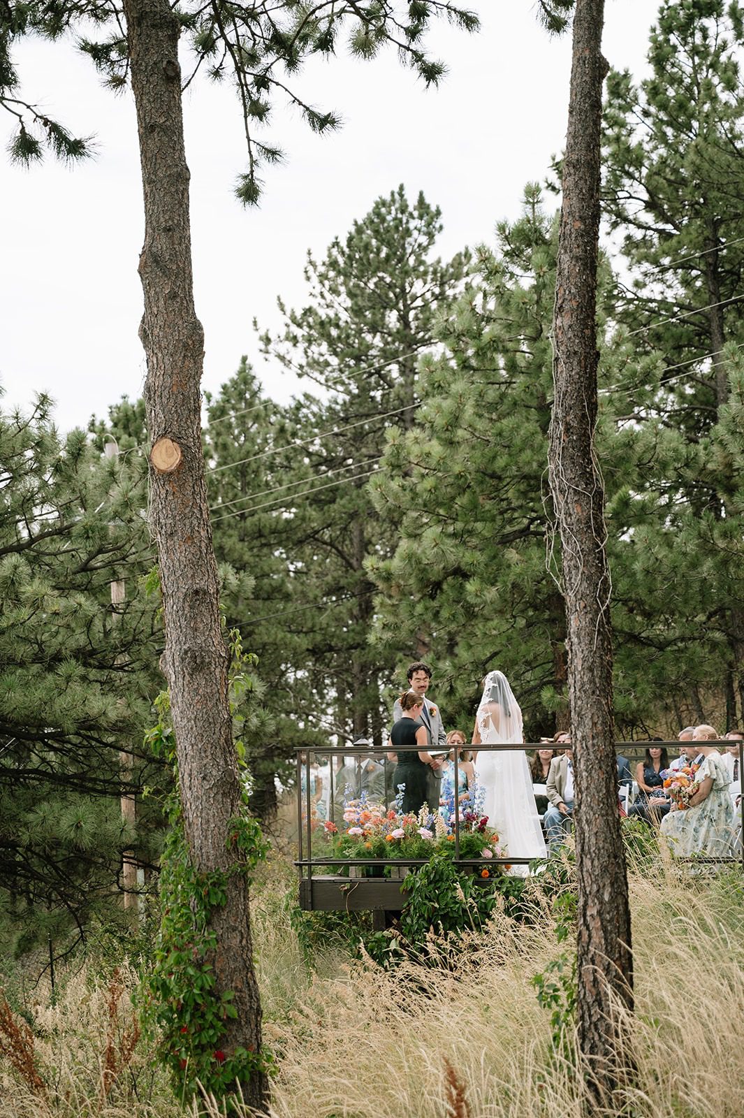 Bride and groom exchange vows on an outdoor terrace framed by tall pine trees, colorful flowers, and seated guests.
