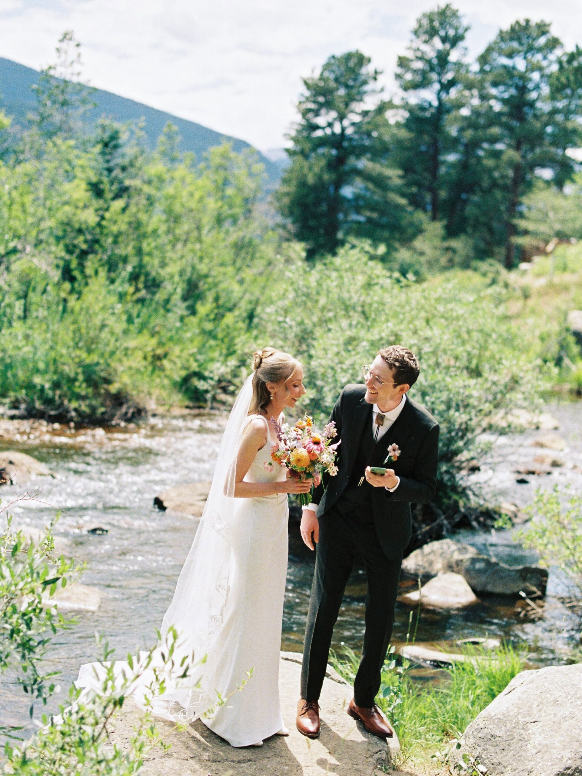 Bride and groom laugh together on rocks beside the river during their Wild Basin Lodge wedding.